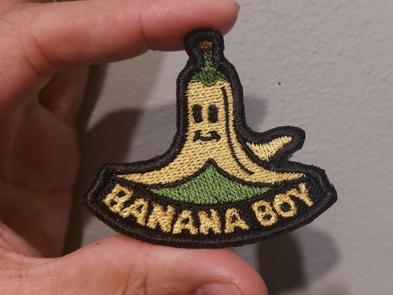 May include: An embroidered patch featuring a cartoon banana character with a yellow peel and green stem. The character has a simple face with two eyes and a mouth. Below the character, the text BANANA BOY is stitched in yellow on a black background.