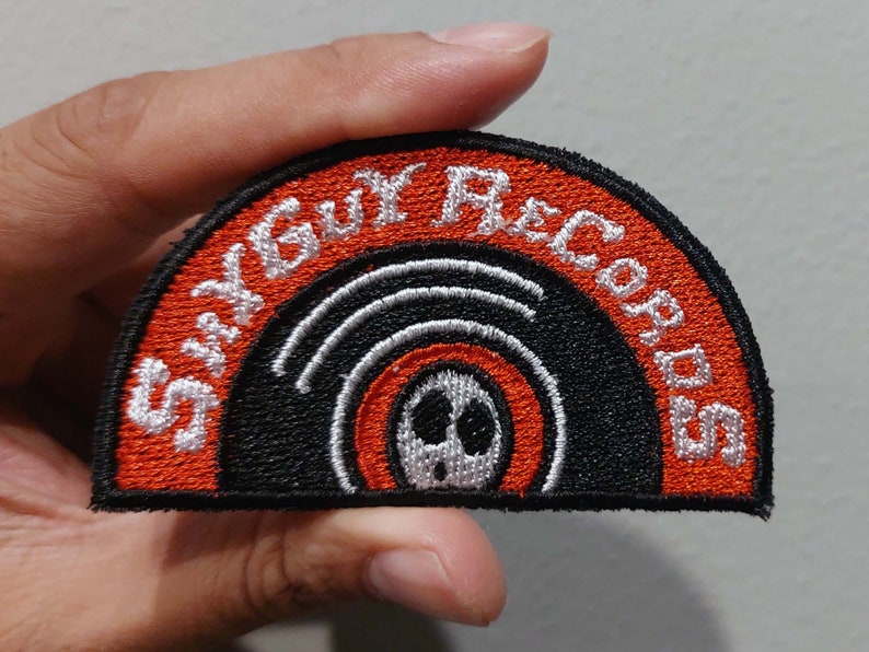 May include: A semi-circular embroidered patch featuring a black and orange vinyl record design. The text ShyGuy Records is stitched in white thread along the curved top edge. A small white character face with black eyes and mouth is centered on the record.
