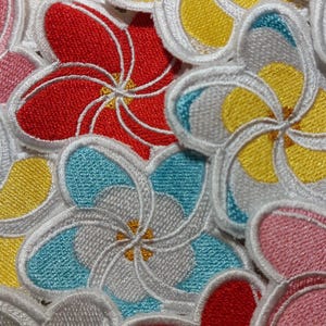 May include: A collection of embroidered flower patches featuring five-petaled blossoms in vibrant colors including red, yellow, pink, and light blue. Each patch has a distinct white stitched border and a small yellow center detail.