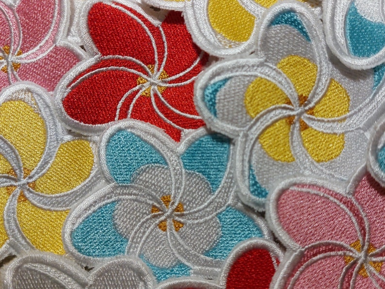 May include: A collection of embroidered flower patches featuring five-petaled blossoms in vibrant colours including red, yellow, pink, and light blue. Each patch has a distinct white stitched border and a small yellow centre detail.