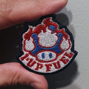 May include: A circular embroidered patch featuring a blue and white mushroom design with red flames at the top. The text 1-UP FUEL is stitched in red capital letters along the bottom edge of the patch, which is outlined with a thick black border.