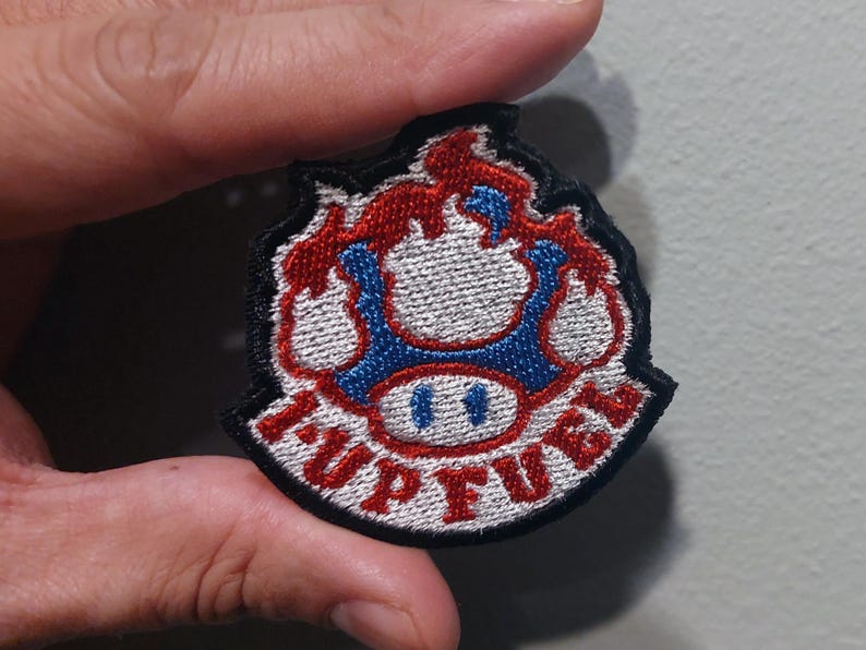May include: A circular embroidered patch featuring a blue and white mushroom design with red flames at the top. The text 1-UP FUEL is stitched in red capital letters along the bottom edge of the patch, which is outlined with a thick black border.