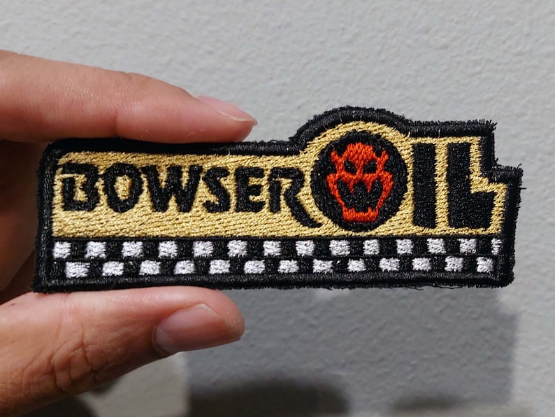May include: A rectangular embroidered patch featuring the text BOWSER OIL in black lettering on a gold background. An orange Bowser emblem replaces the letter O. The bottom section displays a black and white checkered pattern with a black border.