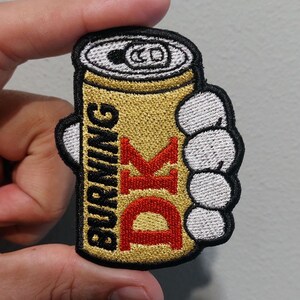 May include: An embroidered patch featuring a gold beverage can with a silver top, held by a white cartoon-style hand. The text BURNING is stitched in black vertically, and the letters DK are stitched in large red font on the gold surface.