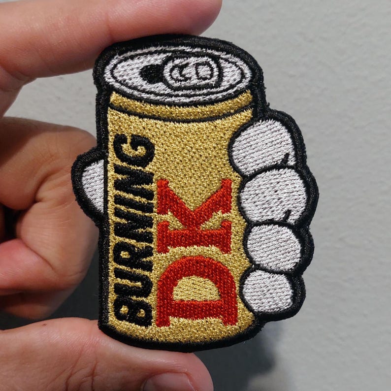 May include: An embroidered patch featuring a gold beverage can with a silver top, held by a white cartoon-style hand. The text BURNING is stitched in black vertically, and the letters DK are stitched in large red font on the gold surface.
