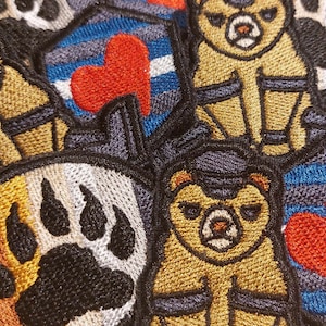 May include: A collection of embroidered patches featuring bears, paw prints, and hearts. The patches are in various colors, including brown, black, white, red, blue, and gray. The patches are made of a soft, textured fabric.