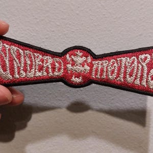 May include: A rectangular embroidered patch with a black border, featuring a red background and silver-colored thread. The text reads "UNDEAD MOTORS" in a stylized font, separated by a central circular emblem with a small cross-like design.