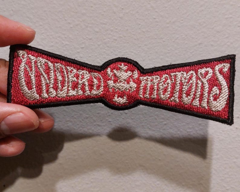 May include: A rectangular embroidered patch with a black border, featuring a red background and silver-colored thread. The text reads "UNDEAD MOTORS" in a stylized font, separated by a central circular emblem with a small cross-like design.