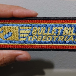 May include: A rectangular embroidered patch with a black border. The design features a blue background with red and gold horizontal stripes. It displays a gold bullet character icon and the text "BULLET BILL SPEEDTRIAL" in gold embroidery.