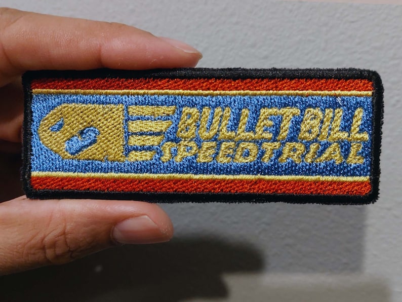 May include: A rectangular embroidered patch with a black border. The design features a blue background with red and gold horizontal stripes. It displays a gold bullet character icon and the text "BULLET BILL SPEEDTRIAL" in gold embroidery.