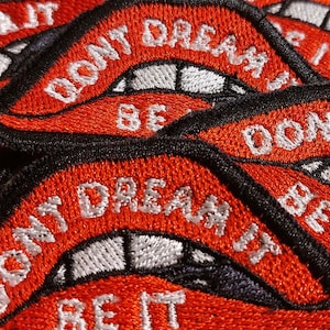 May include: A collection of embroidered patches featuring a red lip design with white teeth and the text DONT DREAM IT BE IT stitched in white thread. Each patch has a thick black border and is layered over others in a repeating pattern.