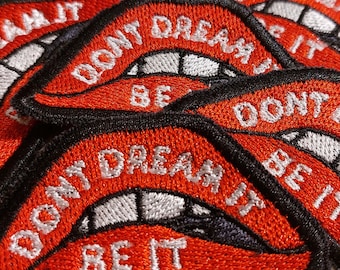 May include: A collection of embroidered patches featuring a red lip design with white teeth and the text DONT DREAM IT BE IT stitched in white thread. Each patch has a thick black border and is layered over others in a repeating pattern.