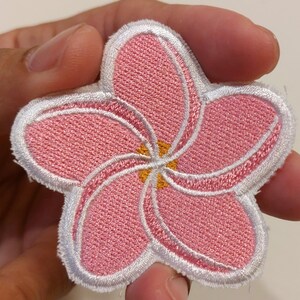 May include: A five-petalled flower patch featuring pink embroidered petals with white stitched borders and a small yellow centre. The design is shaped like a plumeria blossom with a textured fabric surface and a clean white edge outline.