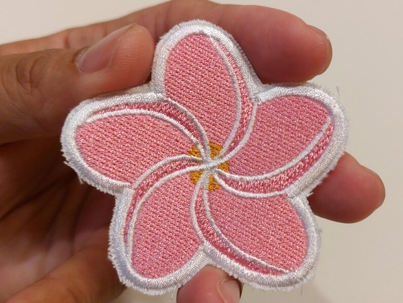 May include: A five-petalled flower patch featuring pink embroidered petals with white stitched borders and a small yellow centre. The design is shaped like a plumeria blossom with a textured fabric surface and a clean white edge outline.