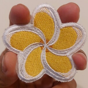 May include: An embroidered patch in the shape of a five-petaled plumeria flower. The petals are bright yellow with a white border and a small orange centre. The stitching is dense and textured, creating a clear floral design with defined edges.