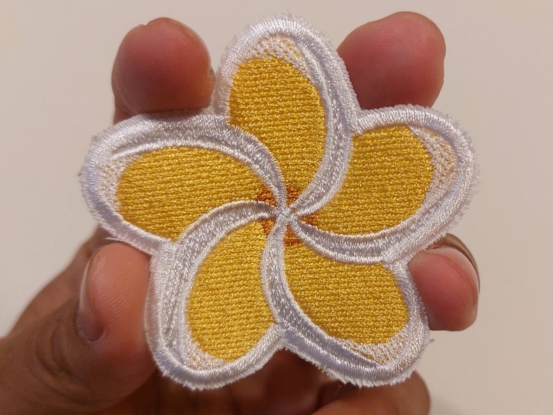 May include: An embroidered patch in the shape of a five-petaled plumeria flower. The petals are bright yellow with a white border and a small orange centre. The stitching is dense and textured, creating a clear floral design with defined edges.