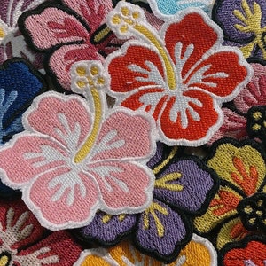 May include: A collection of colorful embroidered hibiscus flower patches. The patches are made of fabric and have a variety of colors, including pink, red, blue, yellow, and purple.