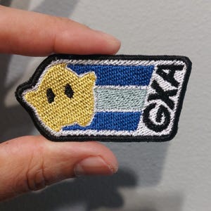 May include: A rectangular embroidered patch with a black border featuring a yellow star with black eyes on the left side. The background consists of horizontal blue and light blue stripes, and the letters GXA are embroidered in black on the right.