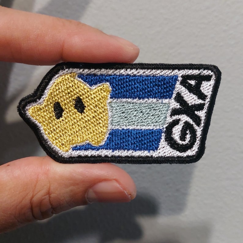 May include: A rectangular embroidered patch with a black border featuring a yellow star with black eyes on the left side. The background consists of horizontal blue and light blue stripes, and the letters GXA are embroidered in black on the right.