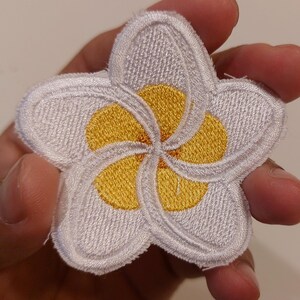 May include: A five-petalled plumeria flower patch featuring white embroidered petals with a bright yellow centre. The design has a distinct stitched border and a textured surface, showcasing a classic floral shape with overlapping petal details.