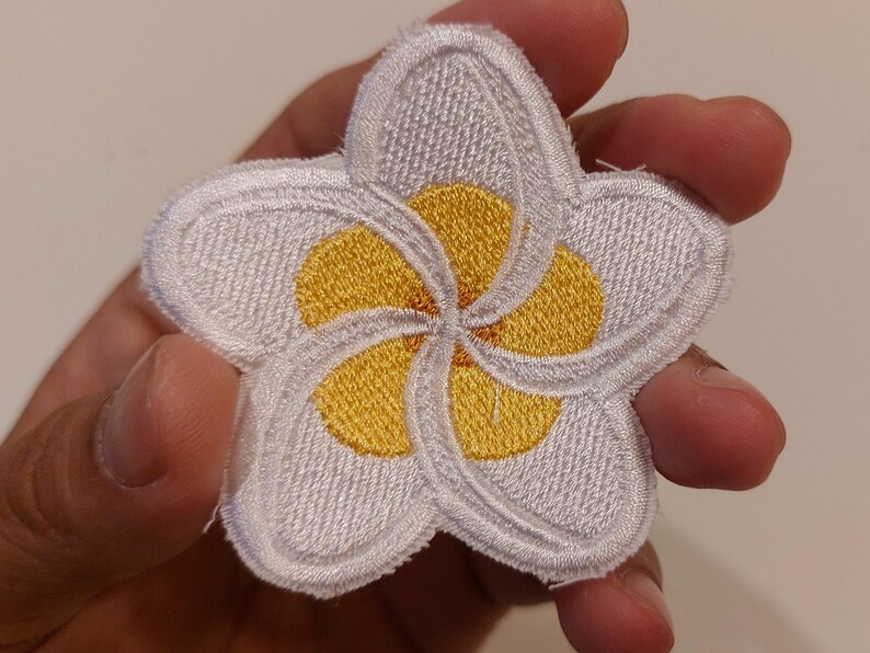 May include: A five-petalled plumeria flower patch featuring white embroidered petals with a bright yellow centre. The design has a distinct stitched border and a textured surface, showcasing a classic floral shape with overlapping petal details.
