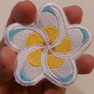 May include: A five-petalled plumeria flower embroidered patch featuring white petals with yellow centres and small light blue accents at the tips. The embroidery has a raised texture and a white stitched border around the entire floral shape.