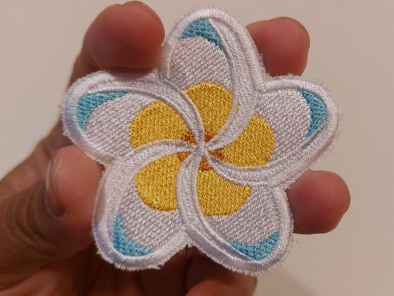May include: A five-petalled plumeria flower embroidered patch featuring white petals with yellow centres and small light blue accents at the tips. The embroidery has a raised texture and a white stitched border around the entire floral shape.