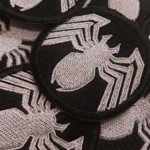 May include: A close-up of several black fabric patches with a white embroidered spider design. The patches are circular and have a stitched edge.