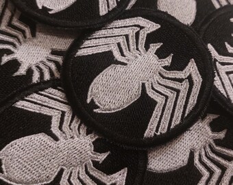 May include: A close-up of several black fabric patches with a white embroidered spider design. The patches are circular and have a stitched edge.