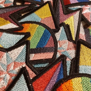 May include: A close-up of several embroidered patches featuring various geometric designs and color combinations, including rainbow, pink, blue, and yellow.