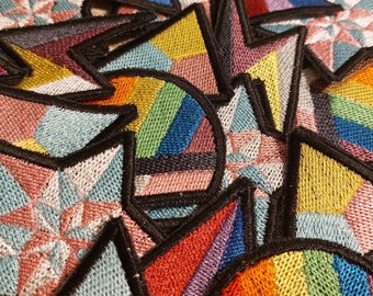 Pode incluir: Um close-up de vários patches bordados com vários desenhos geométricos e combinações de cores, incluindo arco-íris, rosa, azul e amarelo.