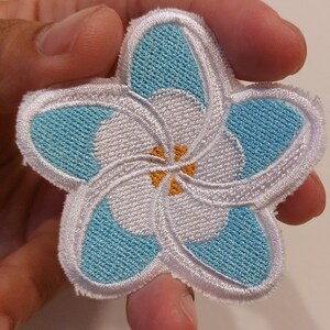 May include: A five-petalled flower patch featuring embroidered light blue and white thread. The design includes a central yellow accent and a white stitched border. The flower shape is symmetrical with overlapping petals in a stylised floral pattern.