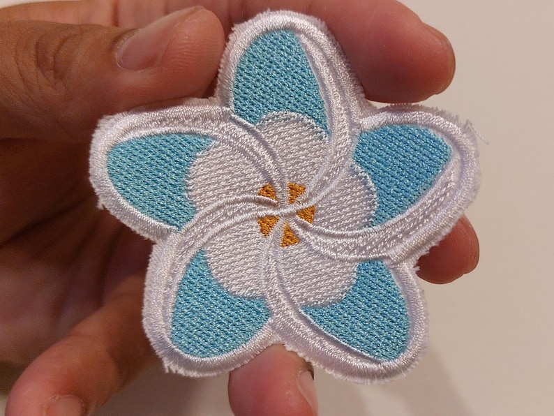 May include: A five-petalled flower patch featuring embroidered light blue and white thread. The design includes a central yellow accent and a white stitched border. The flower shape is symmetrical with overlapping petals in a stylised floral pattern.