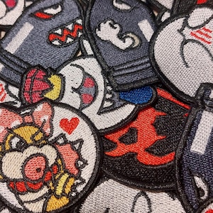 May include: A collection of embroidered fabric patches featuring various cartoon characters and symbols. The designs include a round patch with a character wearing a pink bow, a red heart, a dark blue bullet-shaped character, and red flame motifs.