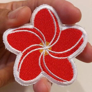 May include: A five-petalled flower patch featuring vibrant red embroidery with white outlines and a small yellow centre. The design is circular with a distinct swirling pattern created by the overlapping red petals and white thread borders.