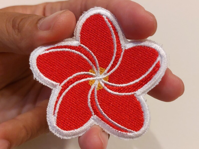 May include: A five-petalled flower patch featuring vibrant red embroidery with white outlines and a small yellow centre. The design is circular with a distinct swirling pattern created by the overlapping red petals and white thread borders.