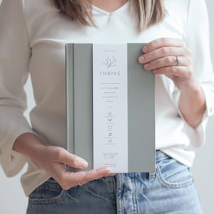 以下が含まれることがあります： 表紙がグレーのハードカバーのジャーナルで、背表紙には「THRIVE」の文字と小さな枝のデザインが施されています。ジャーナルには、アイコンのリストと「Leather Cover」という文字が印刷された白いラベルが付いています。