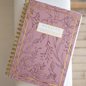 May include: A spiral-bound journal with a mauve cover featuring a floral design and gold accents. The cover has the word "WARRIOR" in a white rectangle. The journal is designed for personal growth and development.