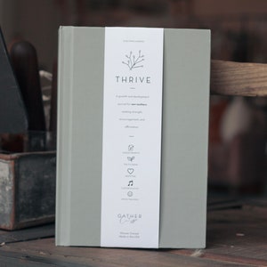 以下が含まれることがあります： 「THRIVE」というタイトルと「新しい母親のための成長と発展のジャーナル。強さ、励まし、肯定を求めています。」というテキストが書かれた灰色のジャーナル。ジャーナルは、小さな枝のデザインと「GATHER Co.」というテキストが書かれた白い背表紙を持っています。