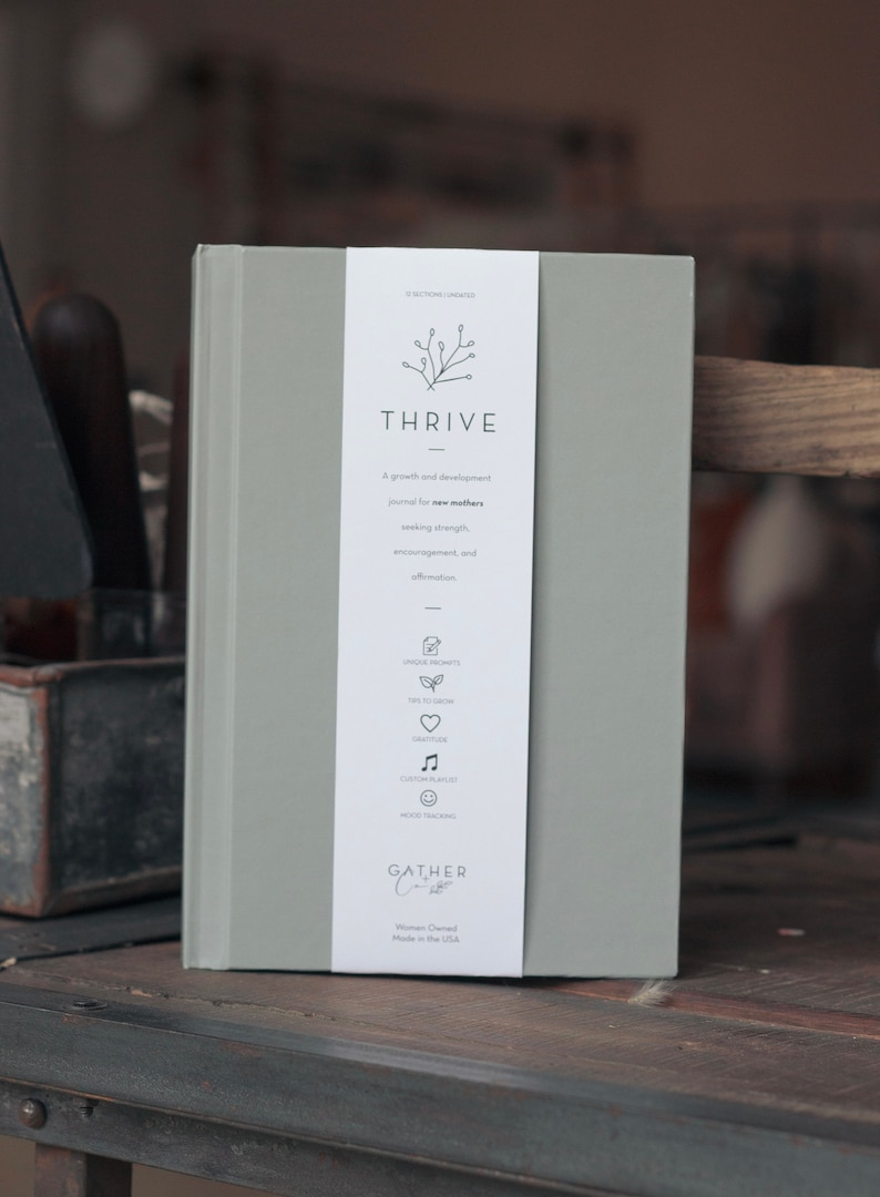 以下が含まれることがあります： 「THRIVE」というタイトルと「新しい母親のための成長と発展のジャーナル。強さ、励まし、肯定を求めています。」というテキストが書かれた灰色のジャーナル。ジャーナルは、小さな枝のデザインと「GATHER Co.」というテキストが書かれた白い背表紙を持っています。
