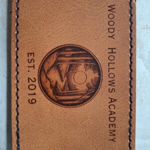 May include: A brown leather patch with a stitched border. The patch features a circular design with a forest scene and a moon. The text "EST. 2019" is on the left and "WOODY HOLLOWS ACADEMY" is on the right.