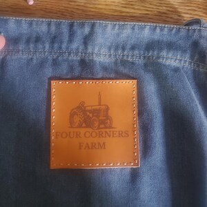 May include: A brown leather patch with a stitched border. The patch features a small tractor illustration and the text "FOUR CORNERS FARM".