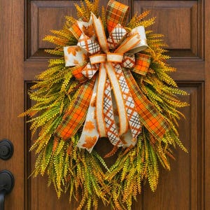 May include: A teardrop-shaped wreath featuring dense, feathery green and yellow foliage. A large, multi-layered bow sits at the top, crafted from ribbons with orange plaid, white and brown checkered, and cream-colored leaf print patterns.