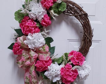May include: A grapevine wreath decorated with pink and white hydrangea flowers and a pink and green patterned ribbon.