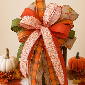 May include: A decorative bow featuring multiple ribbons in autumn colors including orange, green, and tan. The ribbons display patterns of plaid, solid orange, and white with orange leaf prints. The bow is constructed with several loops and long, trailing tails.