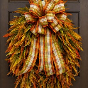 May include: A teardrop-shaped wreath composed of dense, feathery foliage in shades of orange, yellow, and green. A large, multi-looped bow made of plaid ribbon featuring orange, green, and white checks is centered at the top with long streamers.