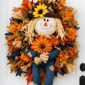 May include: A decorative wreath featuring a plush scarecrow figure with a brown hat, yellow sunflower, and raffia hair. The scarecrow holds an orange pumpkin and wears blue fabric clothing. The wreath is adorned with orange, red, and yellow leaves.