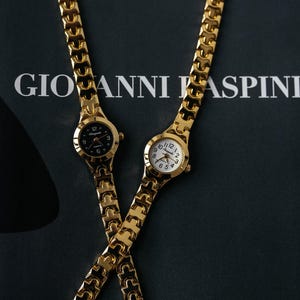 May include: Two gold-tone wristwatches with matching metal bands. The watch on the left has a black face with gold-tone hands and hour markers. The watch on the right has a white face with black hands and hour markers. Both watches feature the brand name "Giovanni Raspini".