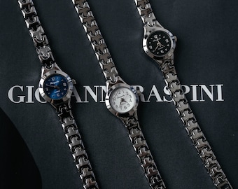 May include: Three silver-toned wristwatches with different colored faces: blue, white, and black. Each watch has a linked metal band. The watches are arranged on a dark surface with the text "GIANNI RASPINI" visible.
