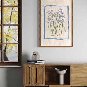 Może przedstawiać: Wydruk w ramie z botanicznym rysunkiem niebieskich kwiatów z zielonymi łodygami na beżowym tle. Na dole wydruku widnieje tekst "Wildflowers for a happier you".